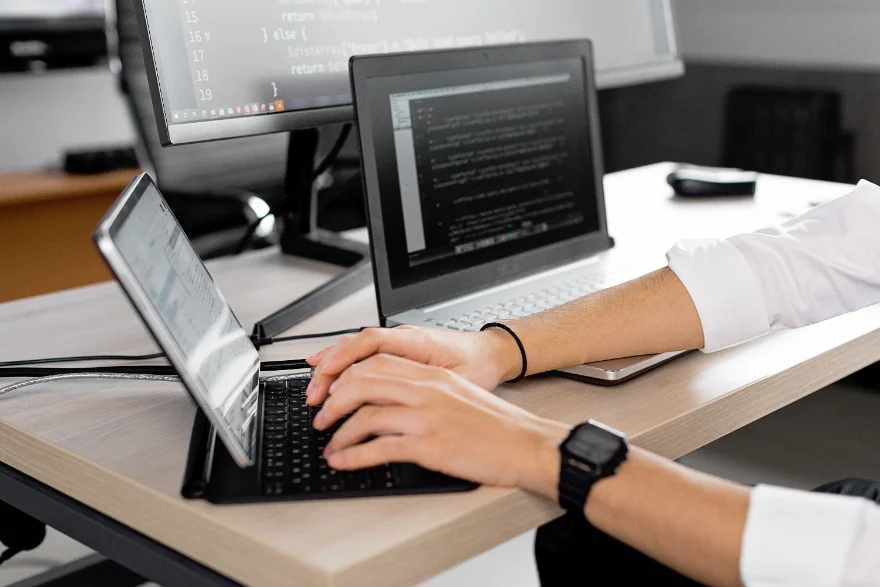 Hands typing on a tablet keyboard at a workstation, with code on a laptop and a monitor alongside